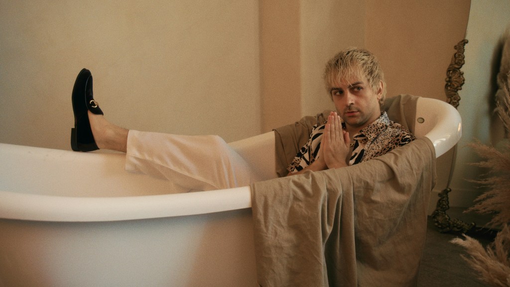 A person with platinum blonde hair sits in a bathtub, wearing a patterned shirt and light trousers, looking thoughtfully while holding their hands together.