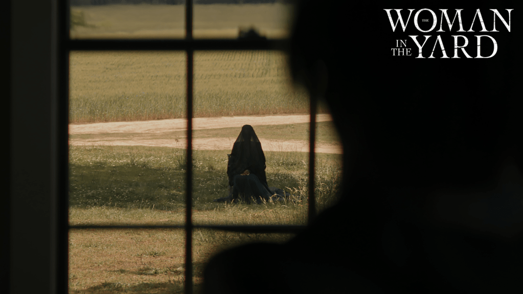 A woman in a dark veil sits in a yard, viewed through a window. The scene is set in a rural area with a hint of tension, emphasizing themes of grief and unease.