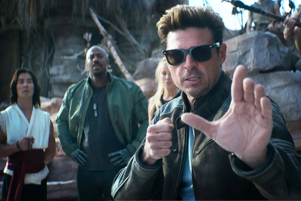 A group of characters in a dramatic scene, with a focus on a man in sunglasses showing a hand gesture, indicating urgency. Other characters are visible in the background, set against a rugged landscape.