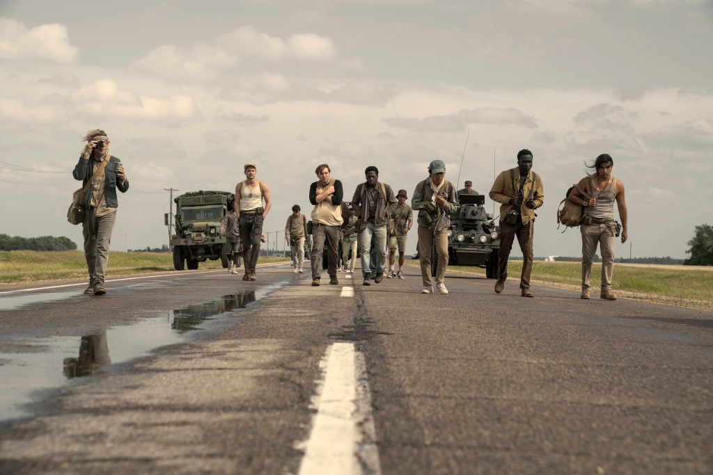 A group of people walking along a highway with military vehicles in the background under a cloudy sky.