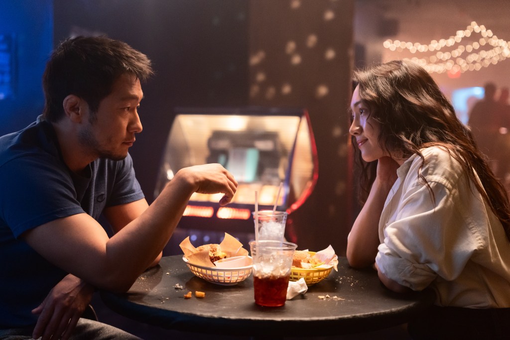 A man and a woman sitting at a table in a dimly lit setting, engaged in a conversation. They have food and drinks in front of them, with a colorful ambiance in the background.